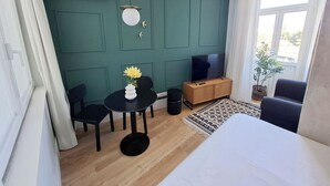 Una habitación de hotel moderna con una pared de acento color turquesa, una mesa redonda con un jarrón de flores amarillas, un televisor sobre un mueble de madera, un sofá negro y un amplio ventanal con vistas.