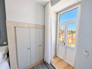 Une chambre à coucher avec un lit, une armoire, une fenêtre donnant sur des immeubles et une porte.