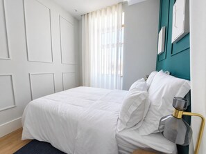Een opgemaakt bed met wit beddengoed in een kamer met teal accentmuren en een raam met transparante gordijnen.