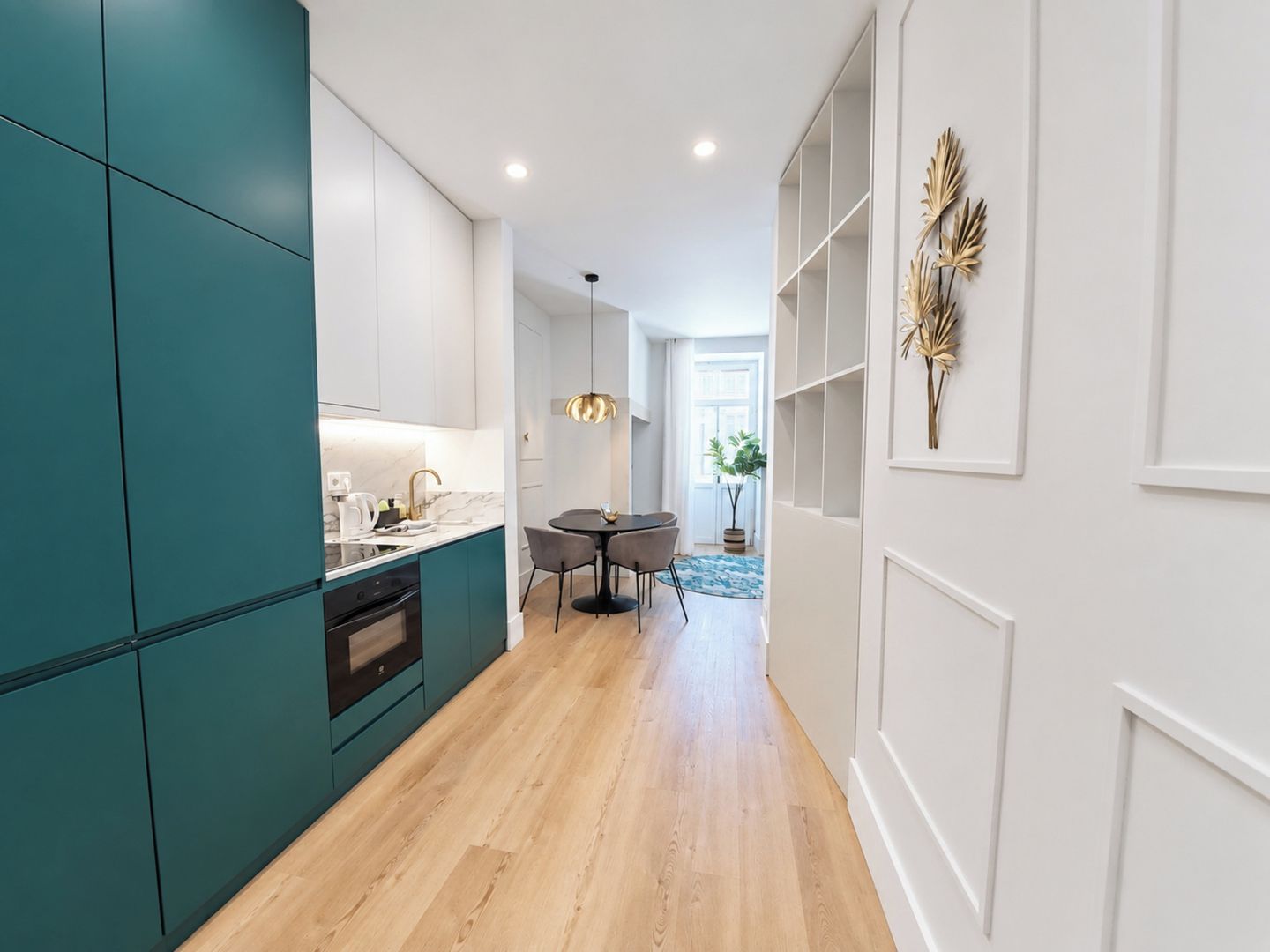 A modern kitchen with teal cabinetry, a dining area with a round table, and a hallway leading to a living space.