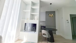 Un salon moderne doté d’un téléviseur encastré, d’étagères blanches, d’un coin repas avec une table et des chaises, et d’un luminaire suspendu.