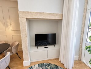 A living room with a fireplace, a flat-screen TV, a dining area with a table and chairs, and a view of the outdoors through a window. - Apartment 105 • Lisbon • 1 bedroom • Elevator • AC (Lisbon)