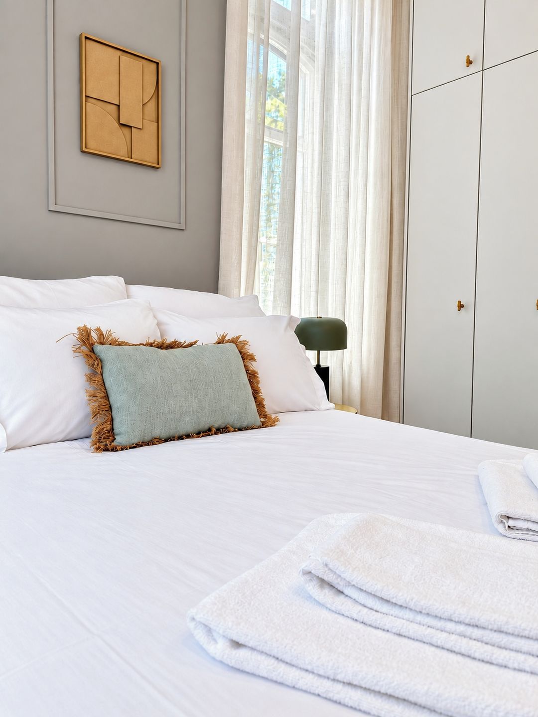 A neatly made bed with white linens and a single decorative pillow, a set of white towels, a framed artwork on the wall, and a wardrobe with a mirror.