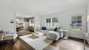 Un salon spacieux avec un canapé blanc, une table basse et un tapis. On aperçoit un coin repas avec une table et des chaises à travers une porte ouverte.