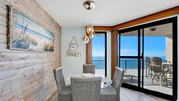 A dining area with wicker chairs and a glass table, a painting of grass and sky, a clock, and a "Beach" sign on the wall. - Beachfront 1BR @ Phoenix X • Sleeps 4 • Gulf Views & Next to Flora-Bama (Orange Beach)
