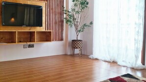 Una sala moderna con piso de madera, un televisor de pantalla plana montado en un panel de madera, una planta en maceta y un ventanal con cortinas translúcidas.