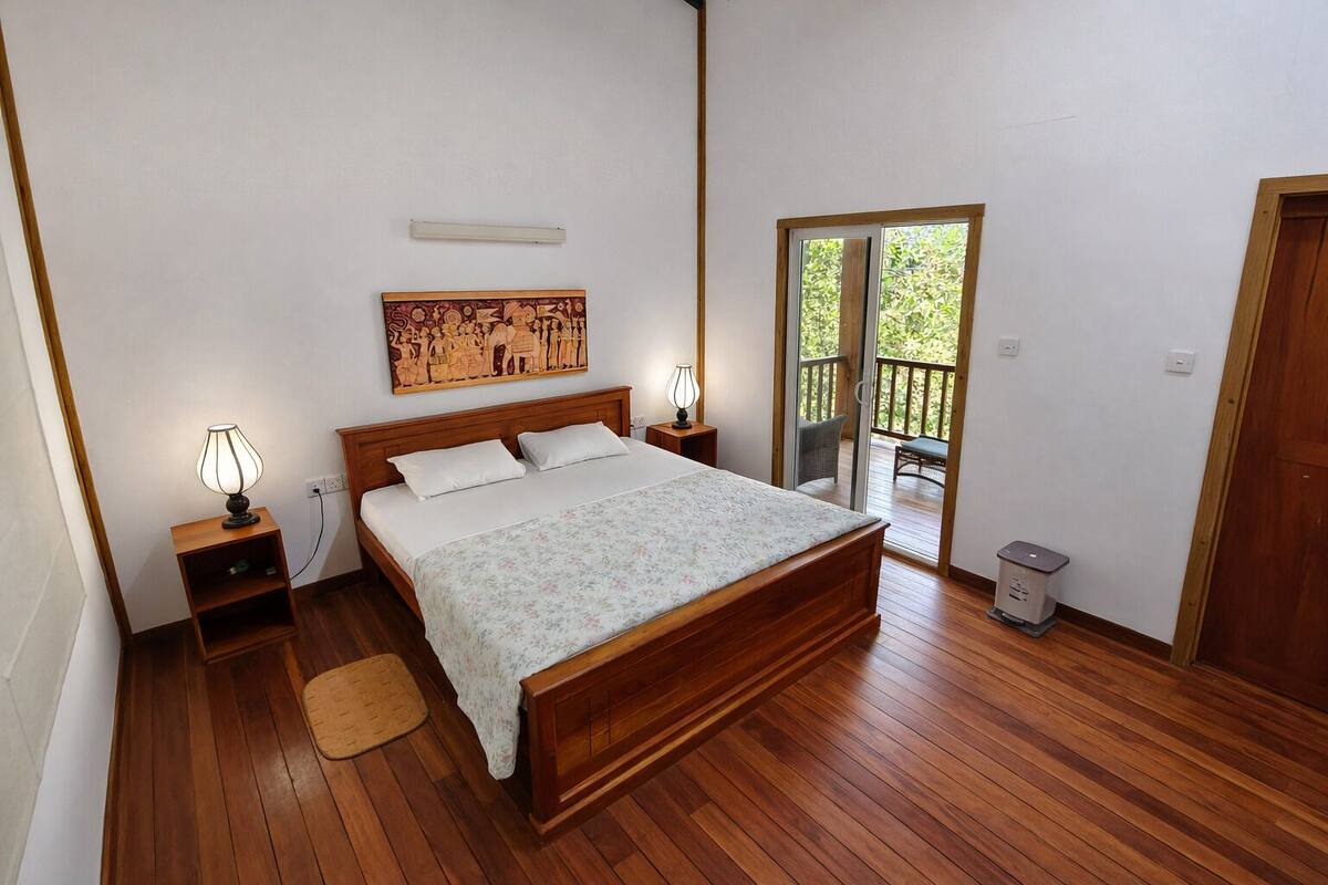 Une chambre à coucher avec un lit en bois, deux lampes de chevet, une table de chevet, un plancher en bois, une porte donnant sur un balcon et un luminaire fixé au mur.
