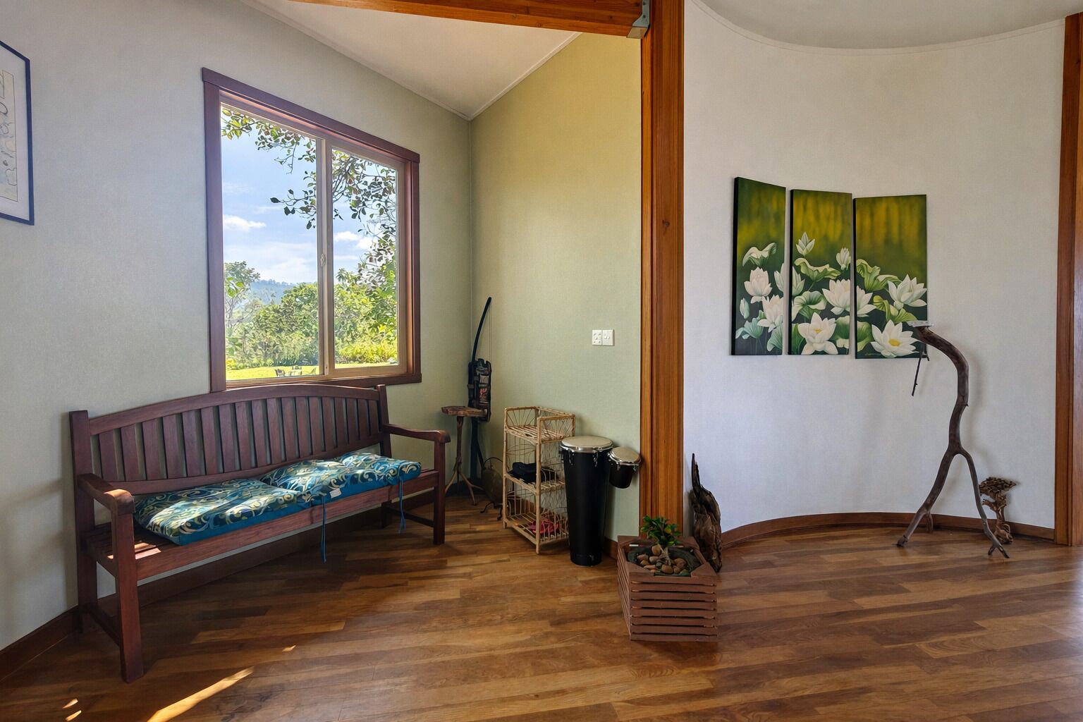 A room with a wooden bench, a large window, a drum set, and a painting of flowers.