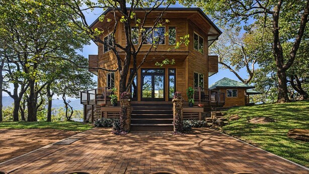 Une maison moderne en bois, dotée d’un balcon, entourée d’arbres et bordée d’un chemin en briques.