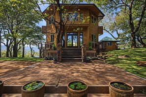 A modern wooden house with a balcony, surrounded by trees and a brick pathway.