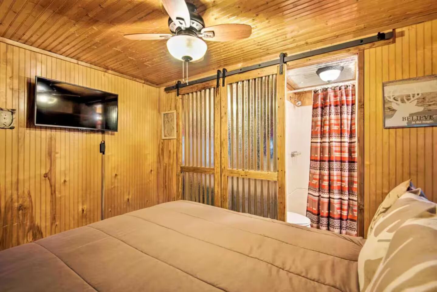 Ett rum med träpanel, en säng, en takfläkt, en tv och en skjutdörr som leder till ett badrum.