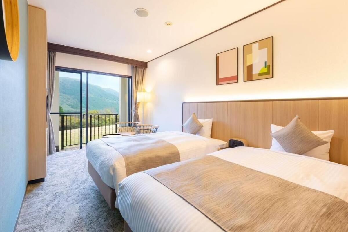 Habitación de hotel con dos camas, un balcón con vistas y un pequeño rincón de descanso.