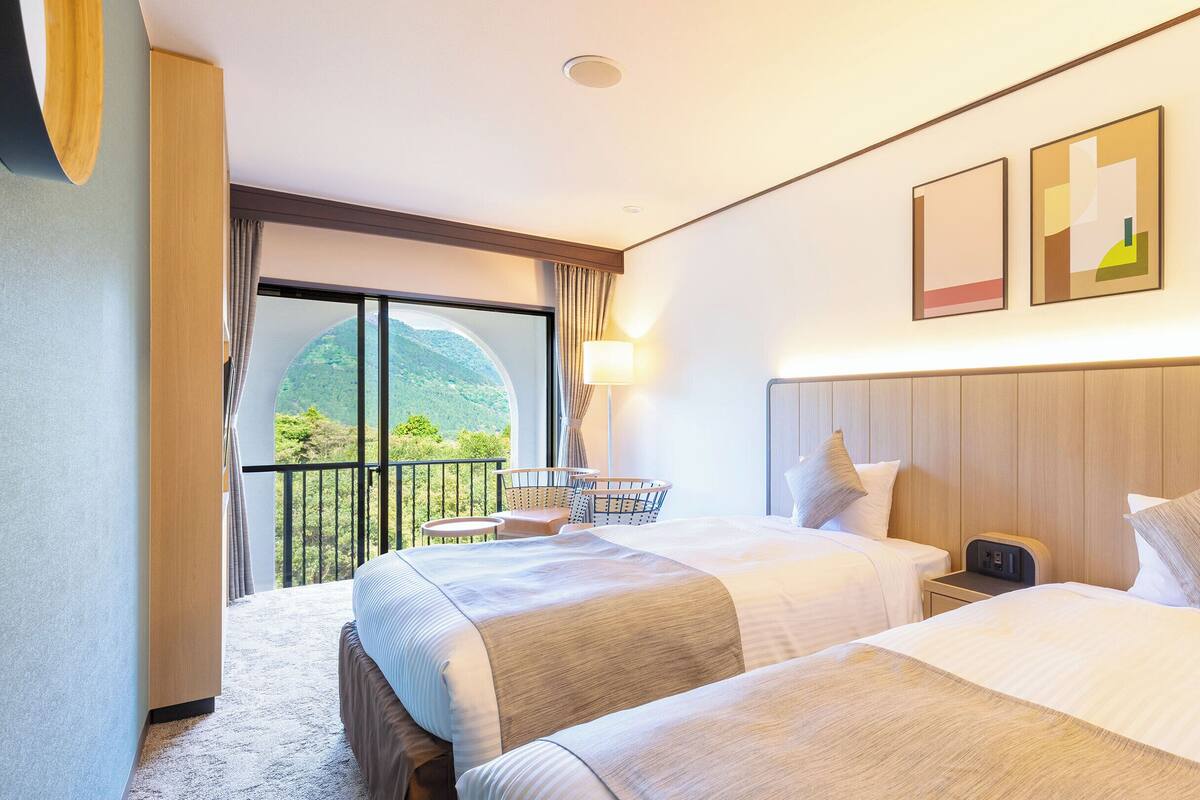 Habitación de hotel con dos camas, un balcón que ofrece vistas a las montañas, una mesita con una silla y una lámpara.