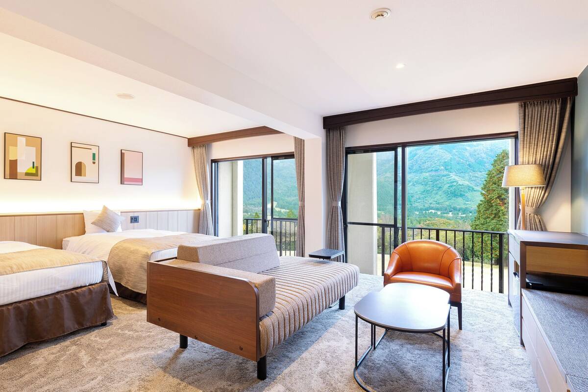 Habitación de hotel con dos camas, un sofá, una silla, un escritorio y un balcón con vistas a las montañas.