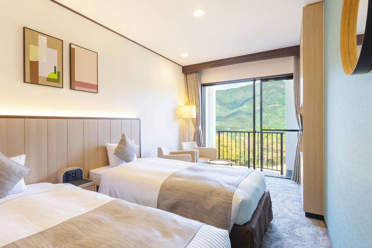 Habitación de hotel con dos camas, una mesita de noche, un escritorio, una silla, una lámpara y un ventanal con vistas a un paisaje montañoso.