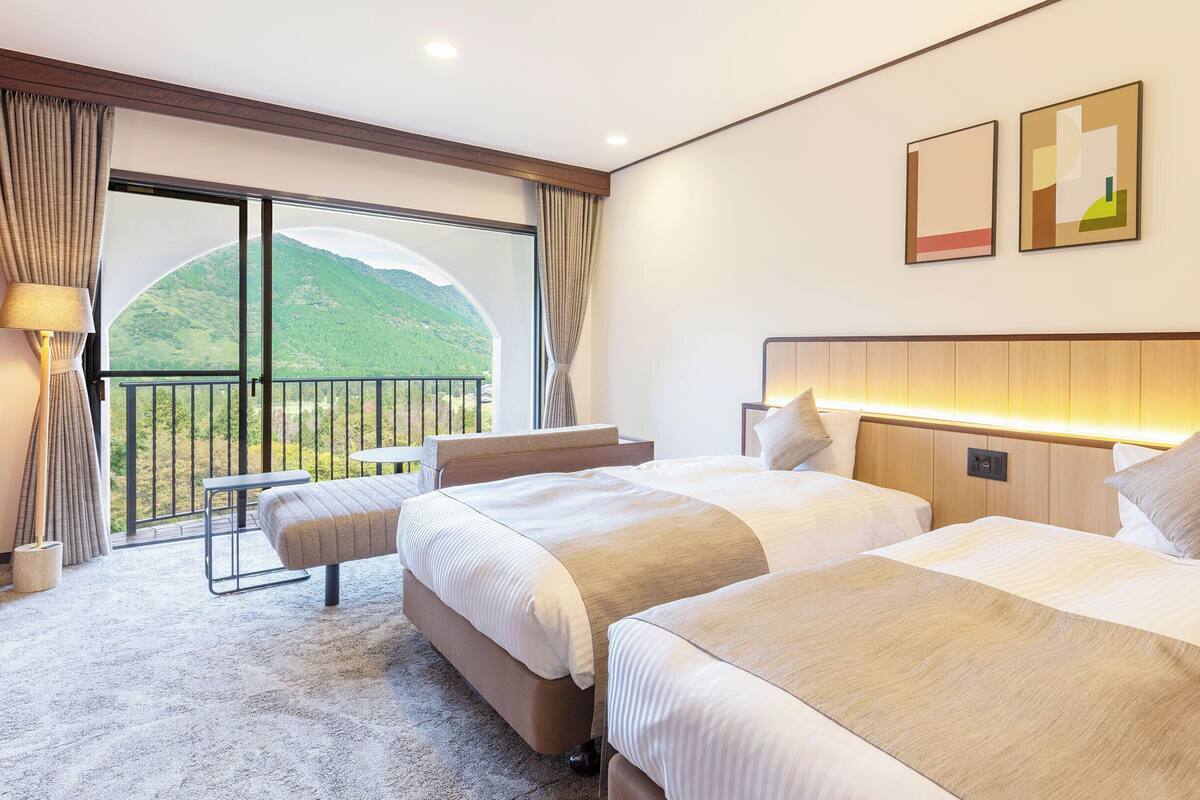 Habitación de hotel con dos camas, balcón y vistas a colinas verdes.