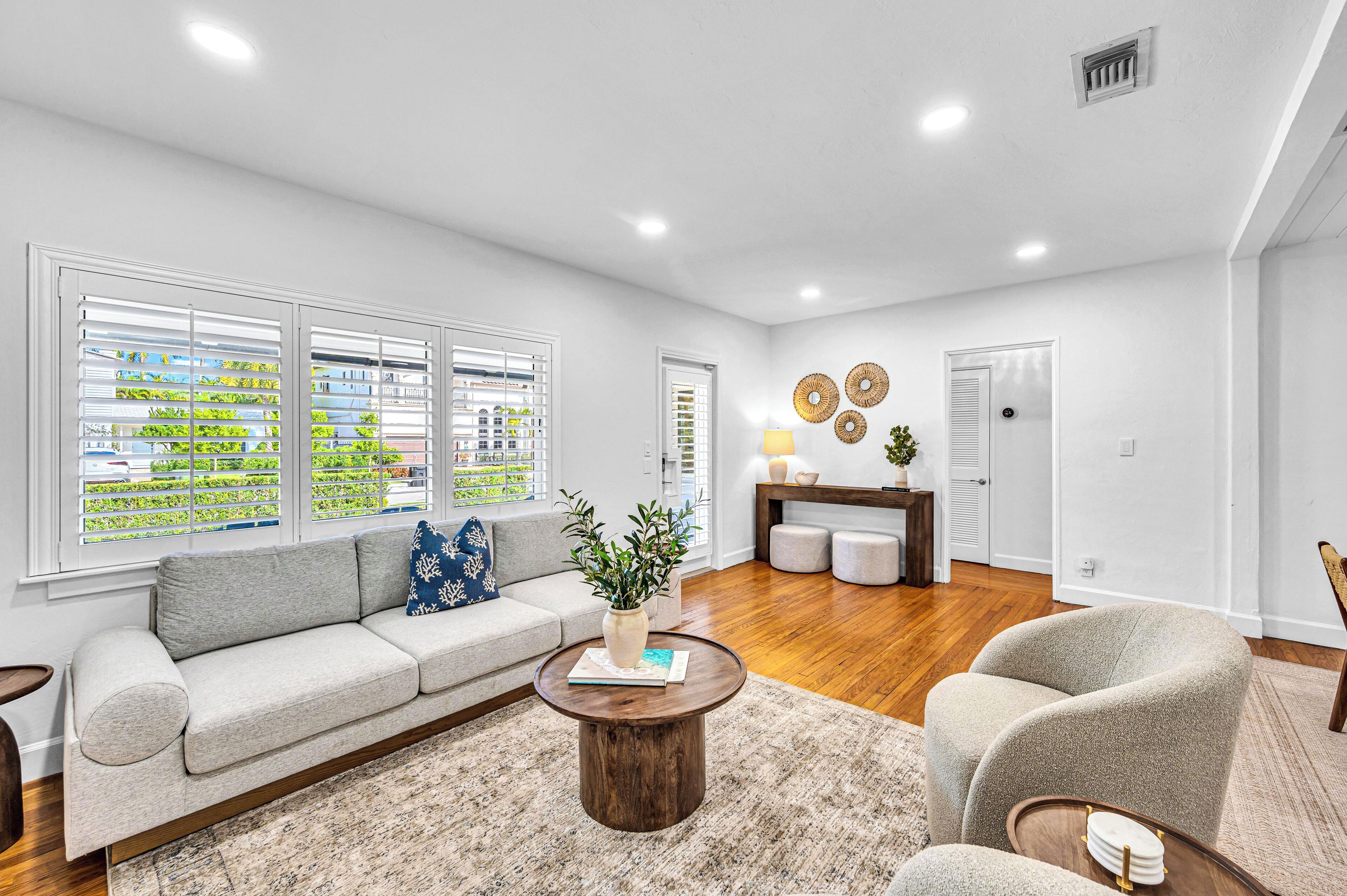 A modern living room with a sofa, coffee table, and a chair. There are windows with blinds, a lamp, and a door.
