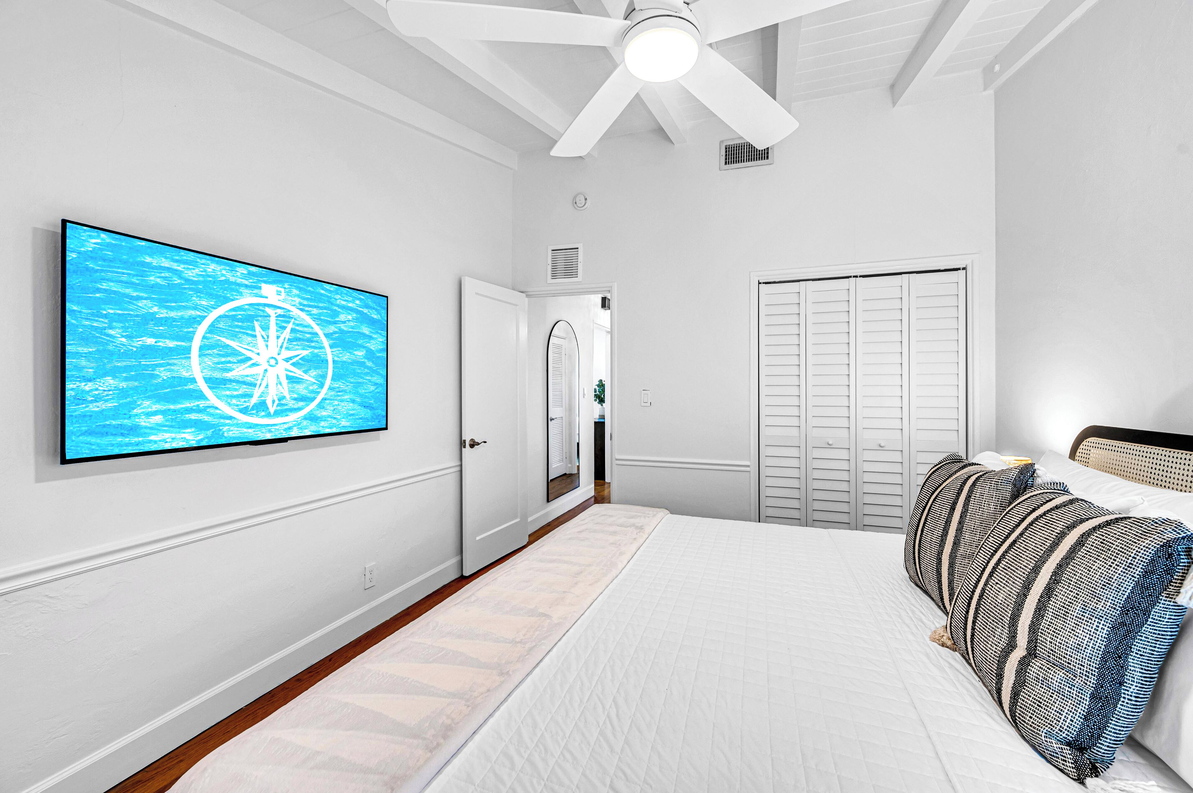 Ein Schlafzimmer mit Bett, Kissen, Deckenventilator, Fernseher, Kleiderschrank und einer Tür zu einem anderen Raum.
