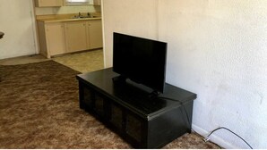 A room with a television on a stand, a kitchen area in the background, and a carpeted floor.
