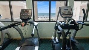 Een sportschool met loopbanden en een elliptische machine, grote ramen met uitzicht op het strand.