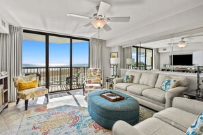 A spacious living room with a large sofa, ottoman, and a chair with a colorful cushion. There is a balcony with a view of the beach and a ceiling fan. - Ocean Bay Club 107 | Oceanfront 4BR Condo | Steps to Main St & Beachfront Pool (North Myrtle Beach)
