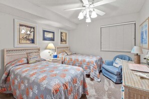 A bedroom with two beds, a ceiling fan, framed artwork on the walls, a window with blinds, and a small table with a lamp. - Ocean Bay Club 107 | Oceanfront 4BR Condo | Steps to Main St & Beachfront Pool (North Myrtle Beach)