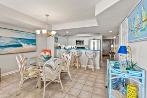 A modern kitchen with white cabinetry, a central island with bar stools, and a dining area with a glass table and wicker chairs. - Ocean Bay Club 107 | Oceanfront 4BR Condo | Steps to Main St & Beachfront Pool (North Myrtle Beach)