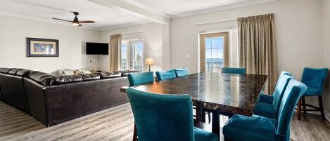 A spacious room with a large wooden dining table surrounded by teal chairs, a leather sofa, and a ceiling fan.