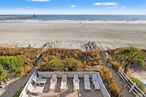 A beach view with a row of white lounge chairs and a wooden deck. - Oceanfront vacation rental with pool, hot tub, and game room—perfect for families and groups (North Myrtle Beach)