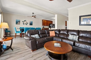 A living room with a leather sectional sofa, a round coffee table, and a dining area with blue chairs. - Oceanfront vacation rental with pool, hot tub, and game room—perfect for families and groups (North Myrtle Beach)