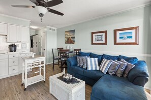 A living room with a blue sofa, white coffee table, and a dining area with a table and chairs. - Direct oceanfront luxury on the 1st floor—step from balcony to beach in seconds (North Myrtle Beach)