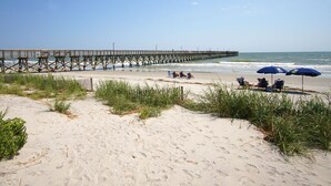 A beach with a wooden pier extending into the ocean, several beach chairs, and umbrellas under a clear sky. - Direct oceanfront luxury on the 1st floor—step from balcony to beach in seconds (North Myrtle Beach)