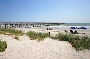 A beach with a wooden pier extending into the ocean, several beach chairs, and umbrellas under a clear sky. - Direct oceanfront luxury on the 1st floor—step from balcony to beach in seconds (North Myrtle Beach)