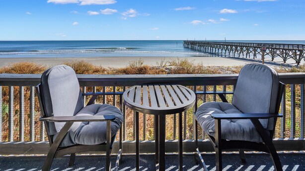 A balcony with two chairs and a small table, overlooking a beach and a pier. - Direct oceanfront luxury on the 1st floor—step from balcony to beach in seconds (North Myrtle Beach)