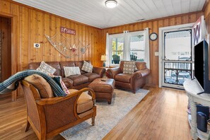 A cozy living room with wooden walls, a leather sofa, a wicker chair, and a television. - Family Beach Retreat with Private Pool, Sunset Views, & Cherry Grove Nature Trails by the Coast (North Myrtle Beach)