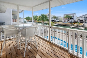 A wooden deck with a glass table and wicker chairs, overlooking a pool and adjacent buildings. - Family Beach Retreat with Private Pool, Sunset Views, & Cherry Grove Nature Trails by the Coast (North Myrtle Beach)