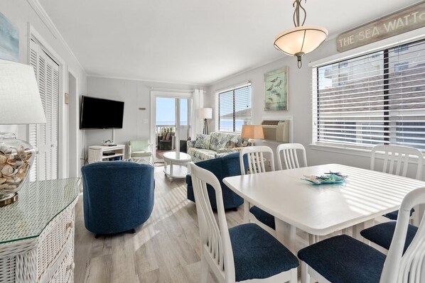 A bright, airy room with a white ceiling, a dining table with chairs, a sofa, and a TV mounted on the wall. - Cozy side-view ocean condo with balcony, pool access, and easy beachside living (North Myrtle Beach)