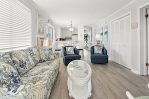 A living room with a floral sofa, two armchairs, a glass coffee table, and a kitchen in the background. - Cozy side-view ocean condo with balcony, pool access, and easy beachside living (North Myrtle Beach)
