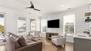 A modern living room with a leather sofa, two striped armchairs, a round coffee table, a flat-screen TV mounted on the wall, and a kitchen area with a sink and countertop. - Exceptional luxury retreat steps from the beach with outdoor oasis, heated pool, elevator (North Myrtle Beach)