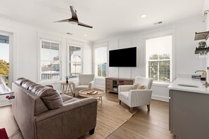 A modern living room with a leather sofa, two striped armchairs, a round coffee table, a flat-screen TV mounted on the wall, and a kitchen area with a sink and countertop. - Exceptional luxury retreat steps from the beach with outdoor oasis, heated pool, elevator (North Myrtle Beach)