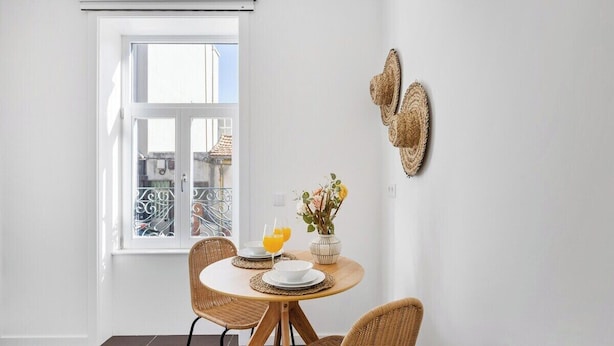 A dining area with a round table, wicker chairs, a vase with flowers, and a window with a view. - Matosinhos Coastal Breeze Studio (Matosinhos)