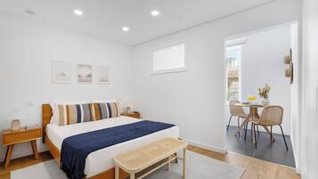 Un dormitorio con cama, mesitas de noche, un banco, un rincón de comedor con sillas y una ventana.