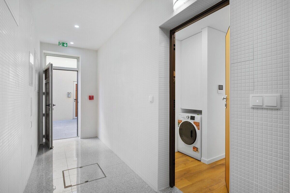 Un pasillo con lavadora, paredes de azulejos blancos y una puerta que da a otra habitación.