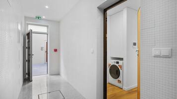 Un pasillo con lavadora, paredes de azulejos blancos y una puerta que da a otra habitación.