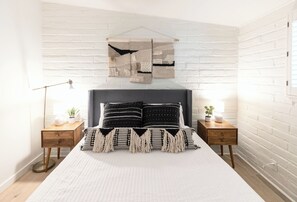 A bedroom with a white brick wall, a bed with black and white bedding, two bedside tables with lamps, and a hanging tapestry. - Tucson Spring Escape | Stylish 3BR w/ Sunny Vibes (Tucson)