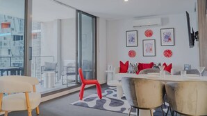 Una sala moderna con puerta de cristal que da a un balcón, un comedor con mesa y sillas blancas, y un sofá con cojines rojos y negros.