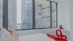 Una habitación de hotel moderna con un ventanal grande, una cama con ropa de cama blanca, una silla roja y un estante pequeño con una taza y un libro titulado 