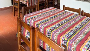 Quarto com duas camas de madeira, roupa de cama colorida, uma mesa de jantar com cadeiras, um ventilador de teto e uma janela com persianas.