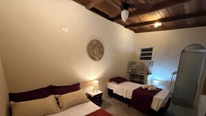 Quarto de hotel com duas camas, uma pequena escrivaninha, espelho, ventoinha no teto e área de chuveiro.