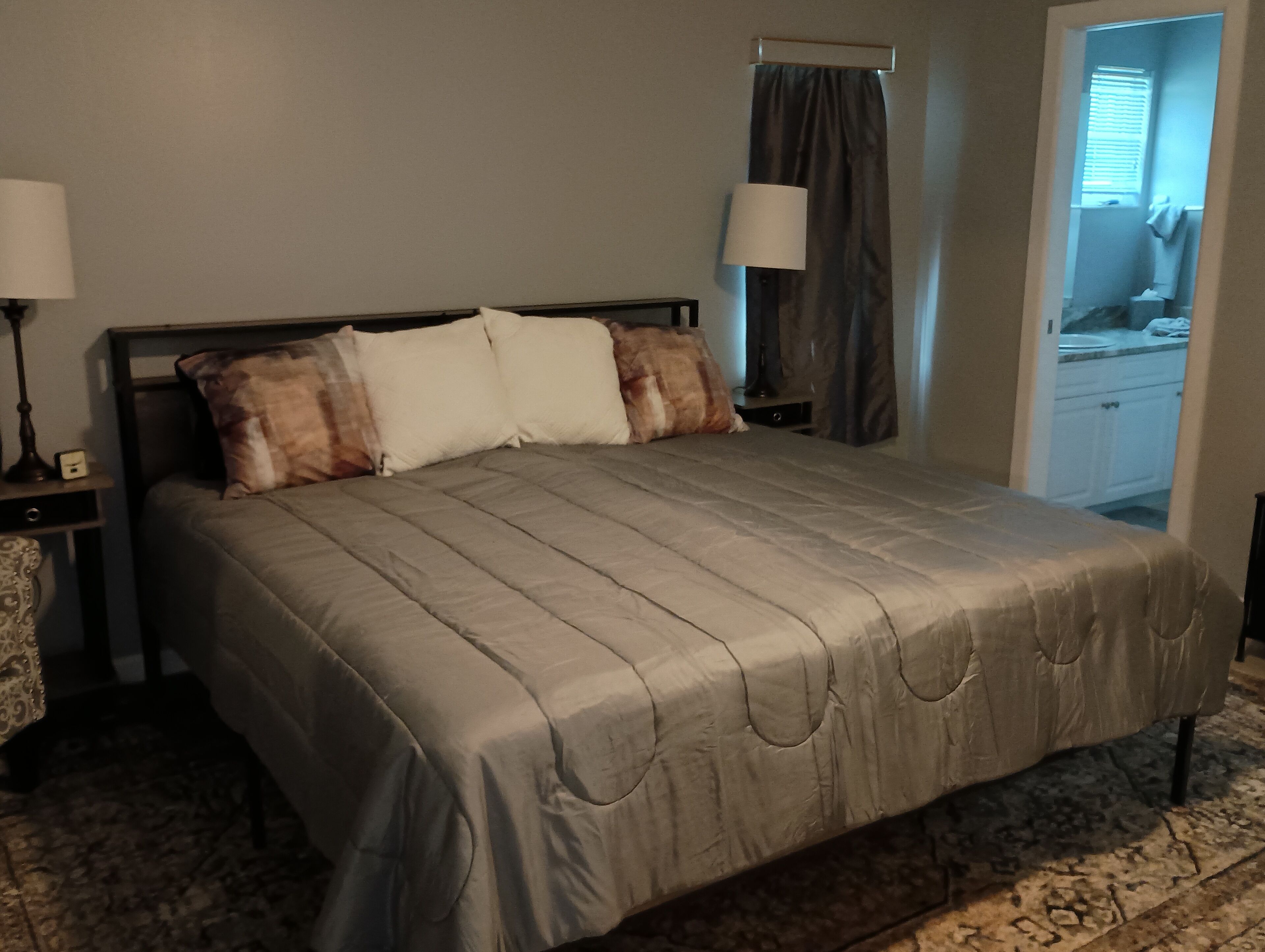 A neatly made bed with a grey bedspread and multiple pillows, a bedside table with a lamp, and a window with curtains.